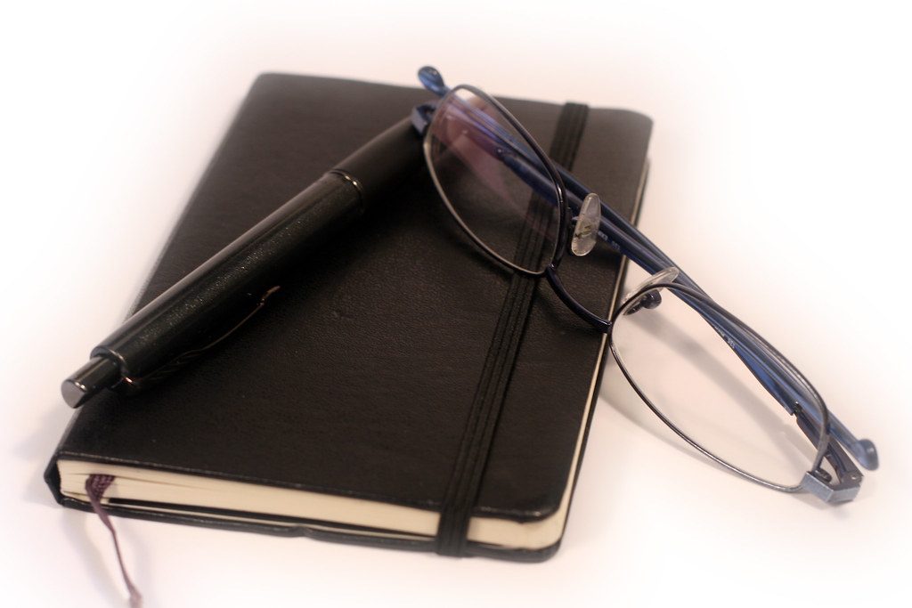 A notebook, a pen and a pair of glasses on a desk